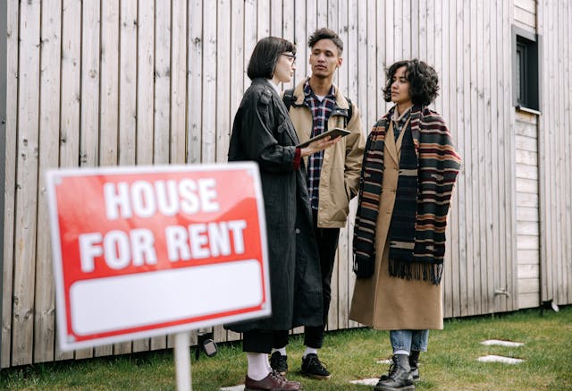 A landlord talking to tenants on a property, behind a “For Rent” sign