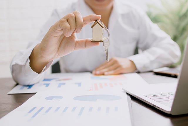 a-person-holding-a-small-wooden-house-keychain-with-a-key-over-a-desk-with-charts-and-a-laptop a-person-holding-a-small-wooden-house-keychain-with-a-key-over-a-desk-with-charts-and-a-laptop
